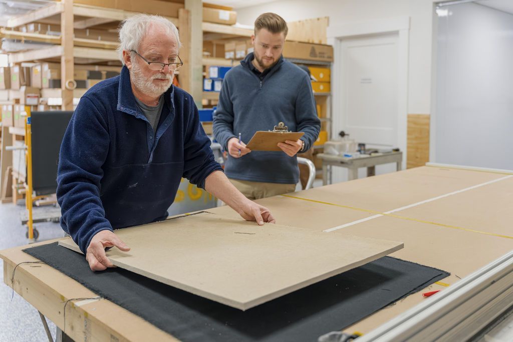 Two workers handling large cardboard sheets in a bright workshop with storage shelves and a cutting table.
