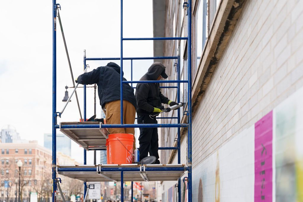 Two workers on scaffolding painting a white brick wall, with an orange bucket and colorful panels nearby