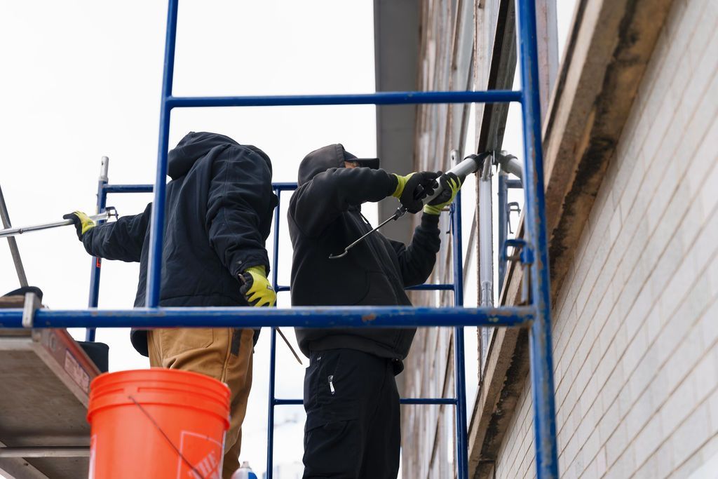 Two workers on scaffolding painting a building exterior beside an orange bucket