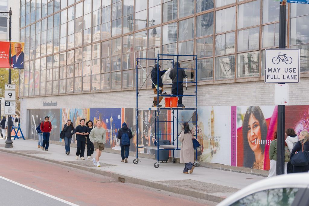People walking past a glass-fronted building with posters along a sidewalk.