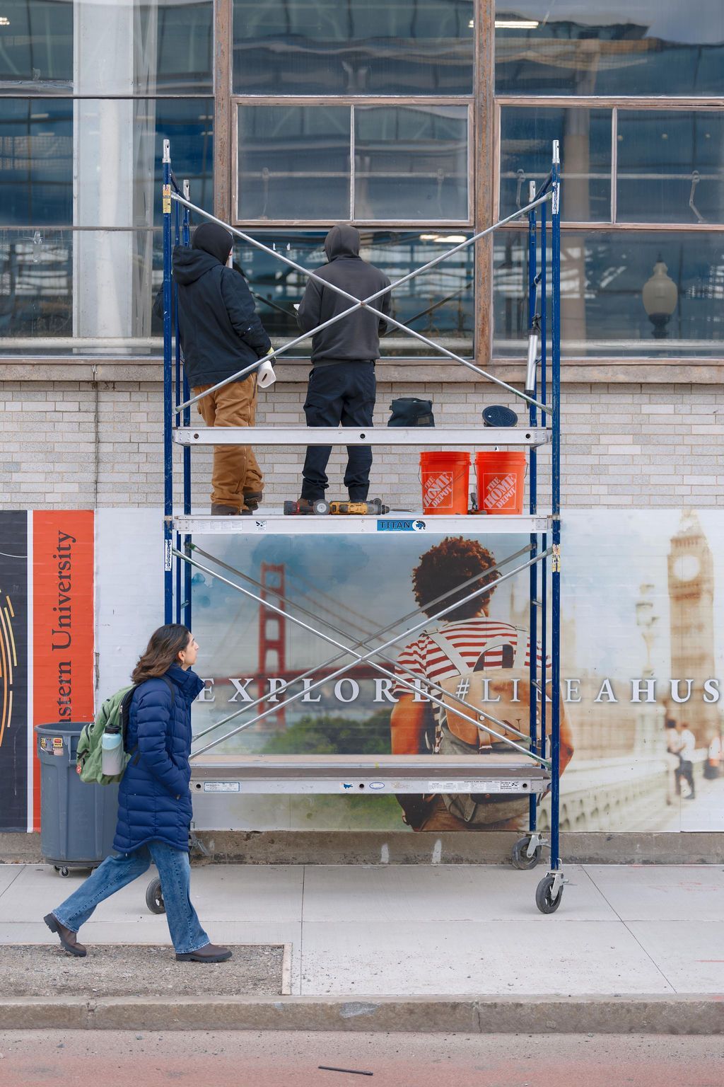 Workers on scaffolding painting a storefront window display on a city sidewalk