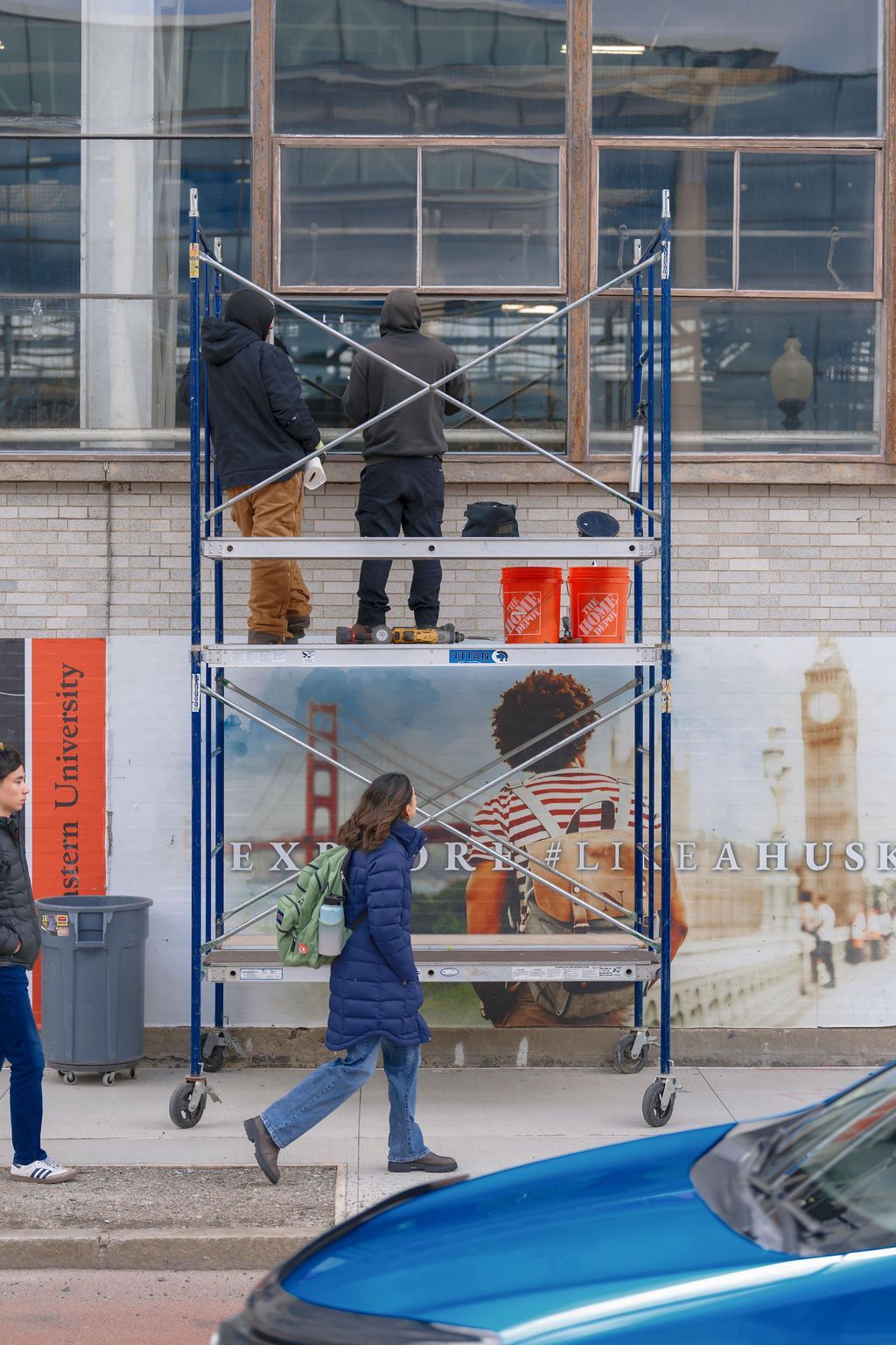 Workers on a scaffold at a building facade, with a blue car passing in front.