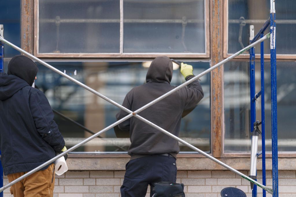 Two workers on scaffolding painting a building’s exterior window frame and wall.
