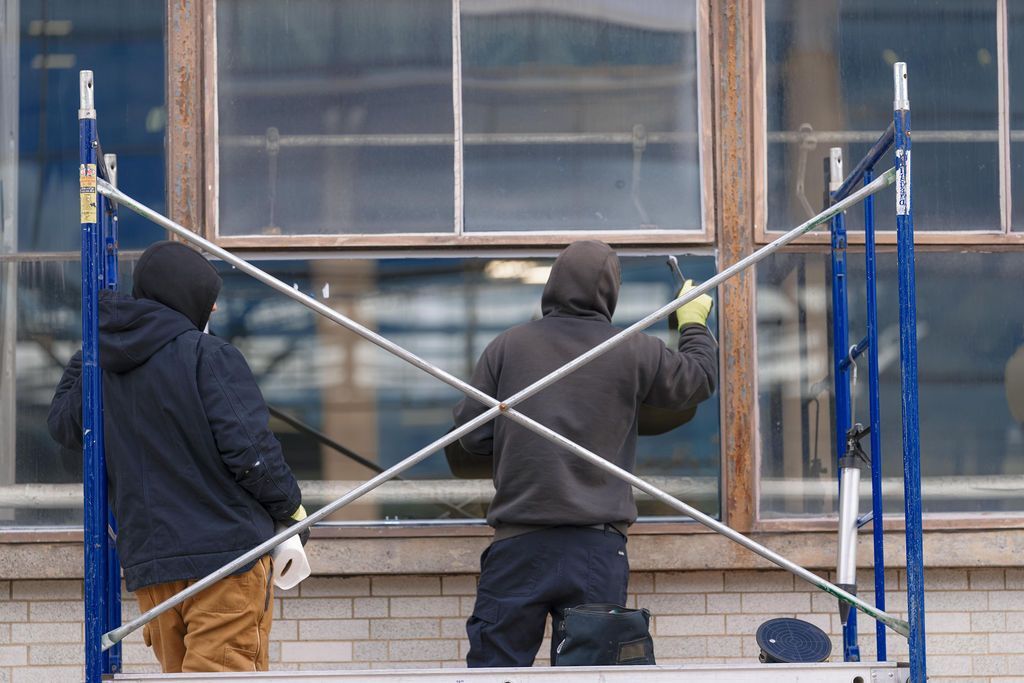 Two workers in hoodies on blue scaffolding cleaning a building window with long poles
