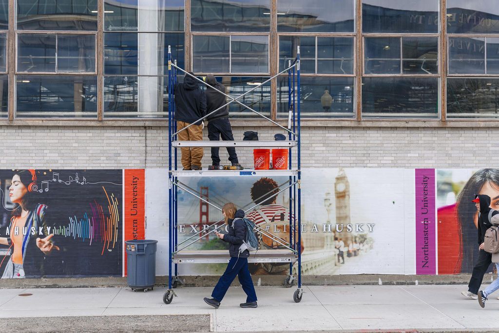 Workers on scaffolding painting a building facade beside a city sidewalk