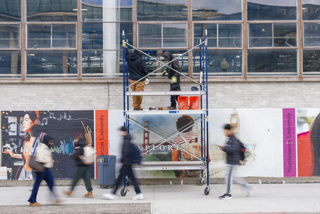 Blurred pedestrians pass a scaffolded storefront with posters and hanging dark blue sculpture.
