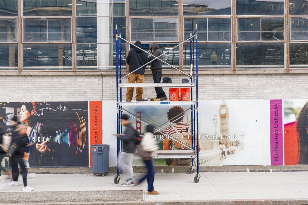 Person on a scaffold painting a wall outside a building, with pedestrians passing by.