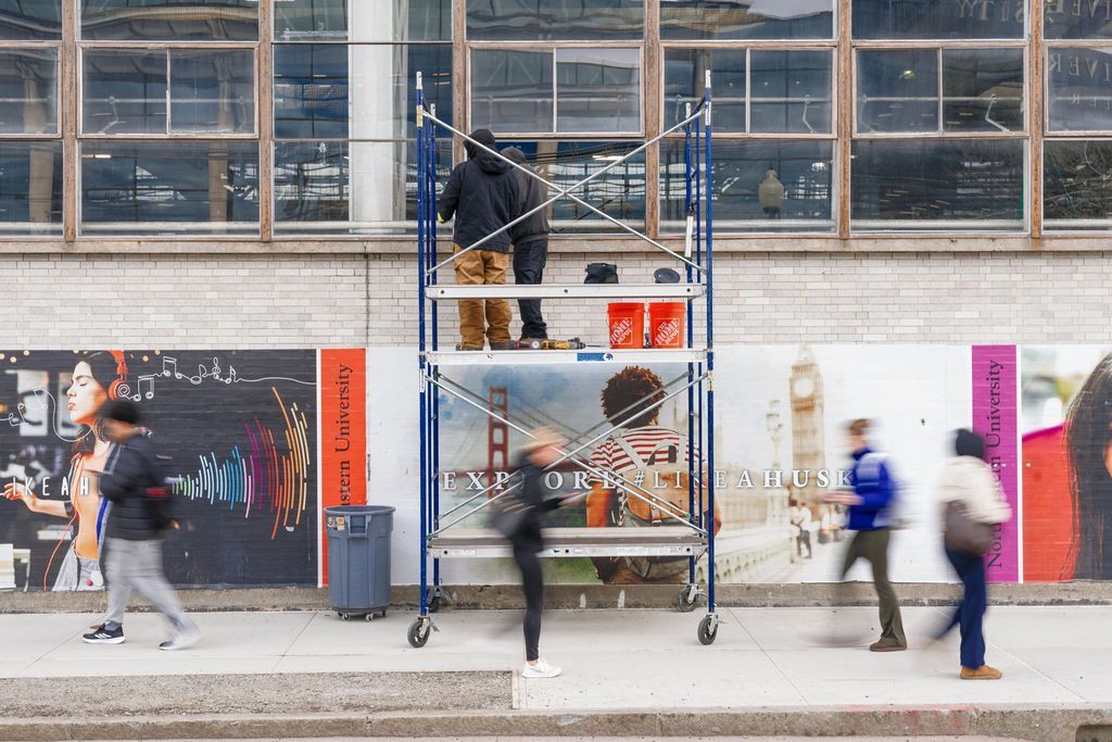 Workers on scaffolding painting a wall outside a building, with pedestrians passing by.