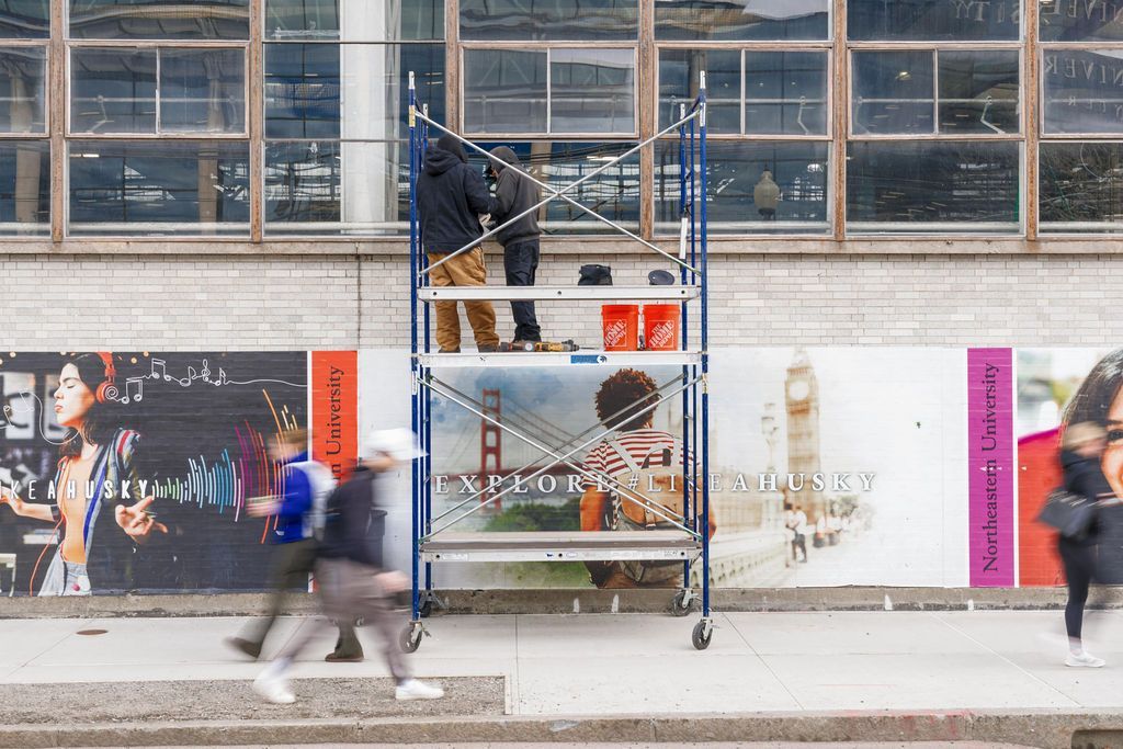 Two workers on a blue scaffold painting a mural on a wall, with a blurred pedestrian passing by.