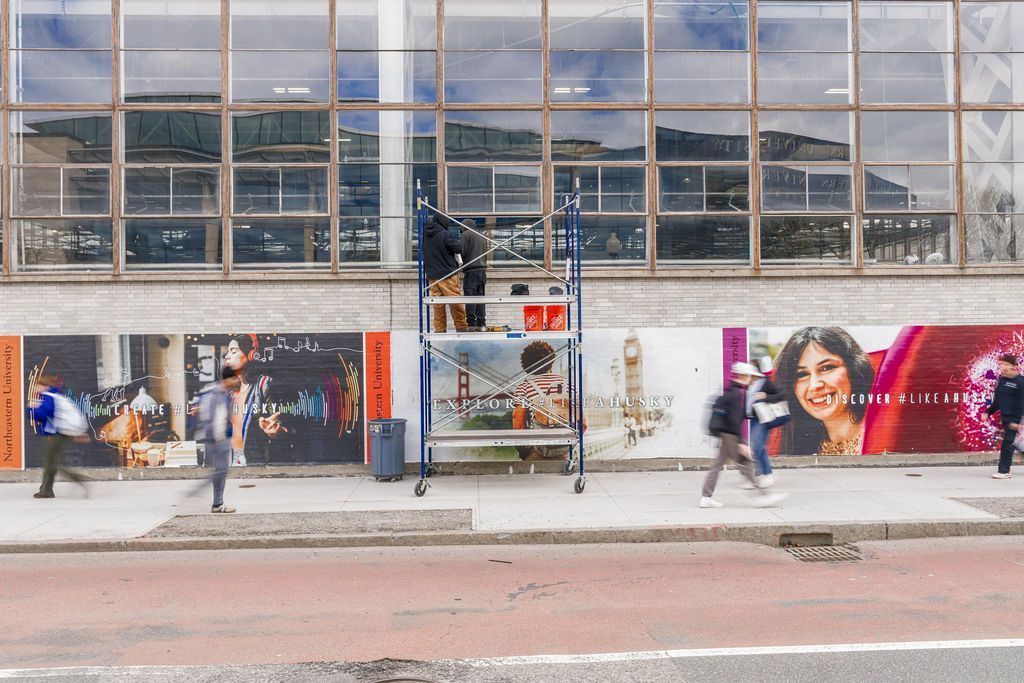 Street storefront with a glass facade, posters, and pedestrians walking past a bus stop in front.