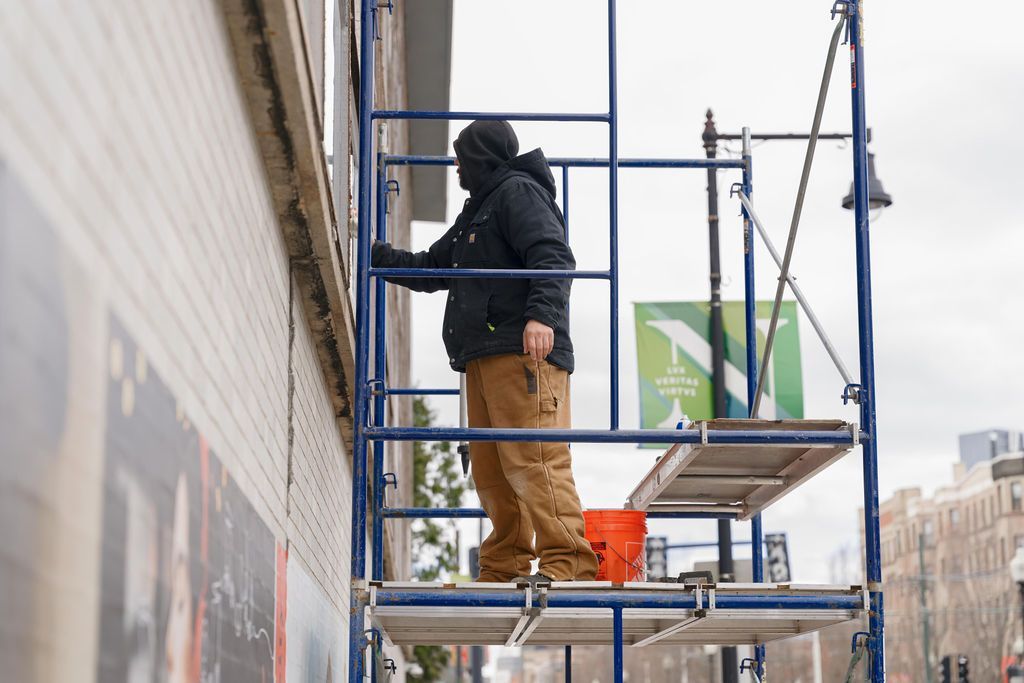 Worker on scaffolding painting a building exterior near a sidewalk, with a bucket and roller.