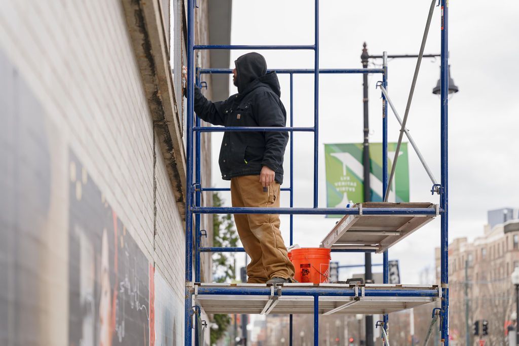 Worker in a black jacket and tan pants on scaffolding beside a wall, painting or working on a building exterior.
