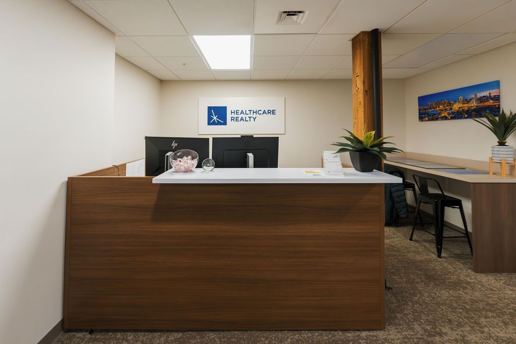 Modern office reception desk with wood front, white countertop, chairs, and wall art in a carpeted lobby
