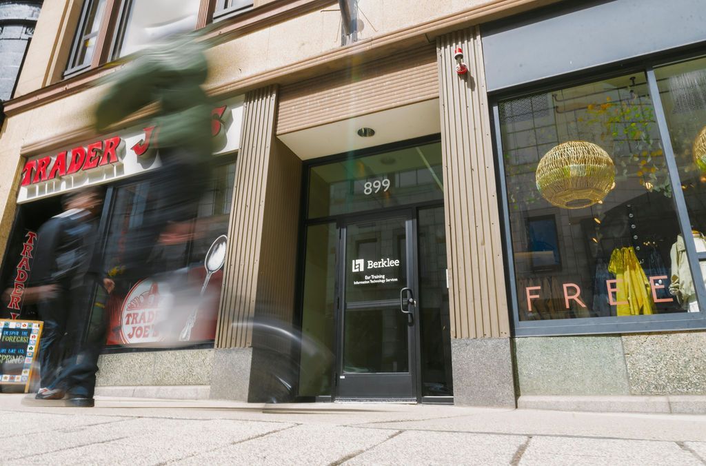 Storefront with a black door and large window displays, seen from the sidewalk as a person passes by blurred.