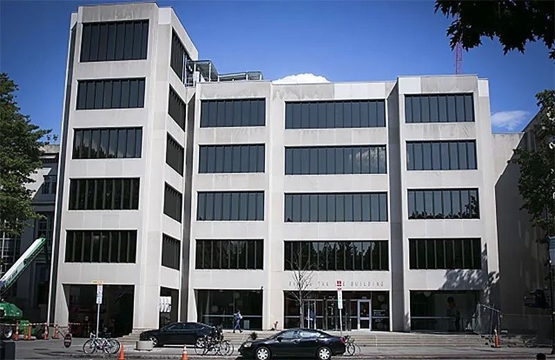 A modern, five-story concrete office building with rows of dark, tinted windows under a clear blue sky.
