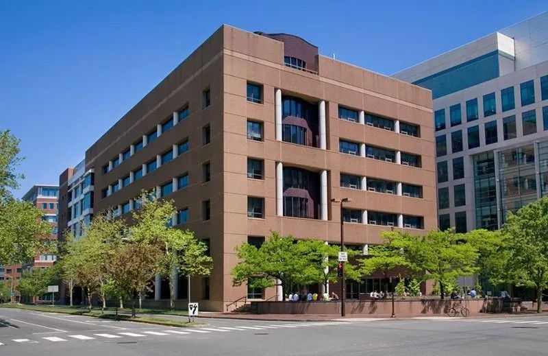 A multi-story brown brick building with modern windows stands on a street corner, surrounded by green trees under a clear sky.