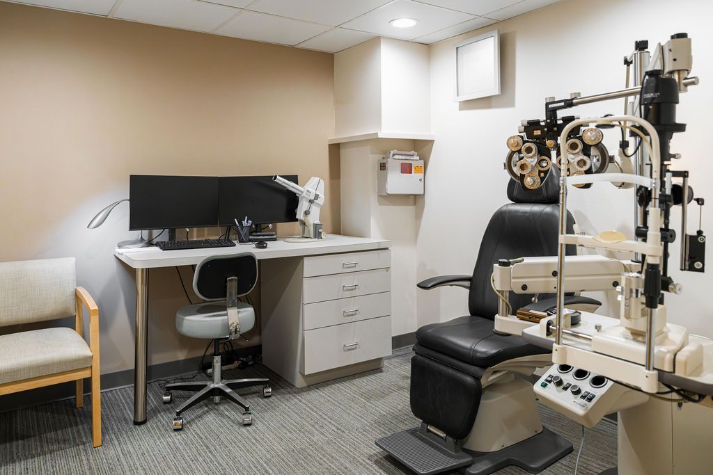 Eye exam room with exam chair, phoropter, desk, computer, and waiting chair.
