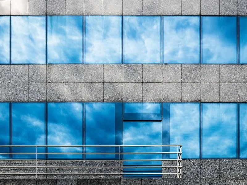 Modern building facade with stone grid pattern and blue glass windows reflecting a cloudy sky, featuring a metal railing.