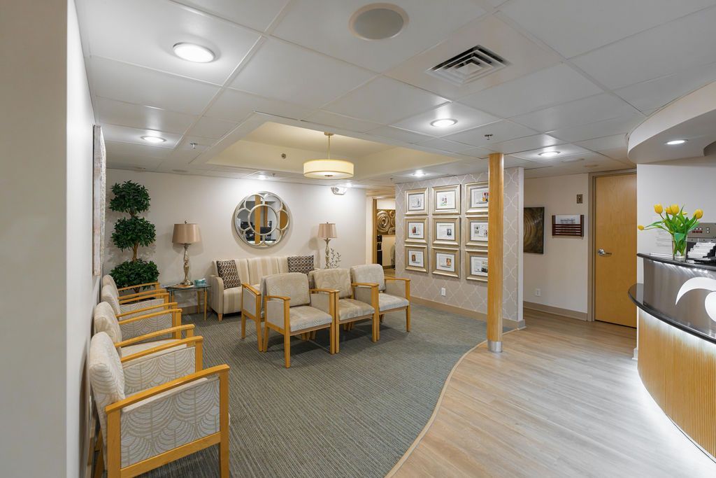 Modern lobby waiting area with wooden chairs, tables, potted plants, and a reception desk