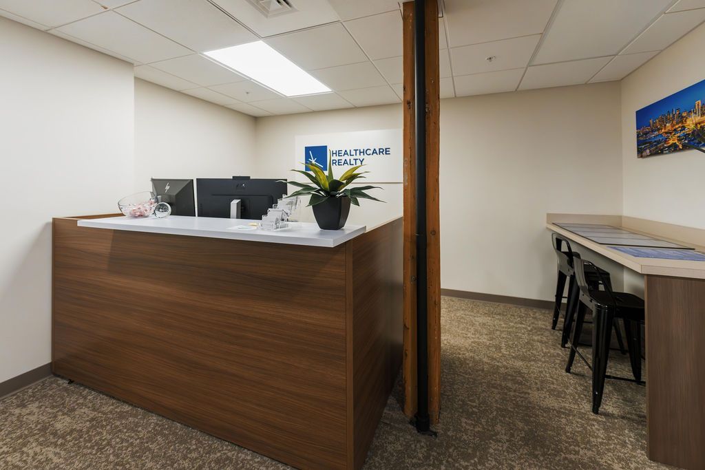 Empty reception area with wood desk, computer monitors, and a narrow table with stools in a beige office corridor