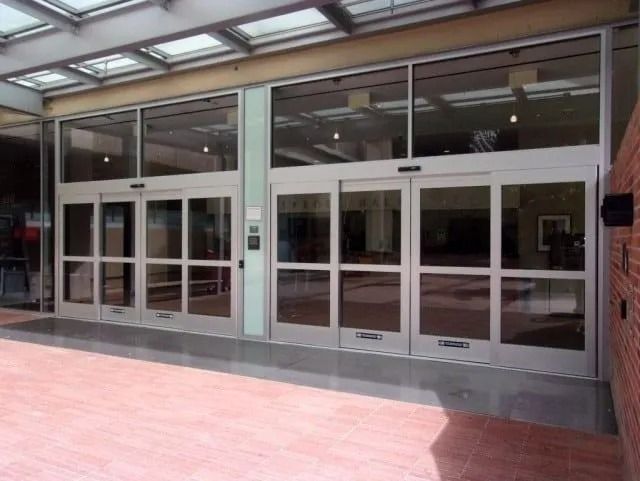 A double set of sliding glass automatic doors at a building entrance with brick siding and a glass canopy.