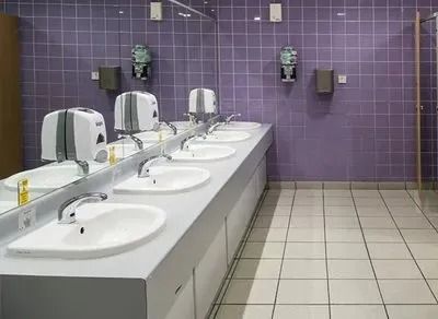 A row of white sinks with soap dispensers against a purple tiled wall in a clean public restroom.