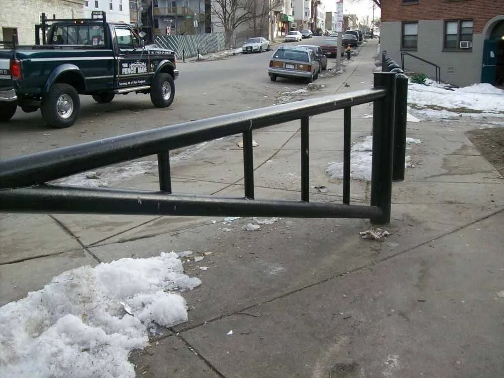 A black metal bike rack sits on a concrete sidewalk next to a road with a parked pickup truck and a grey sedan.