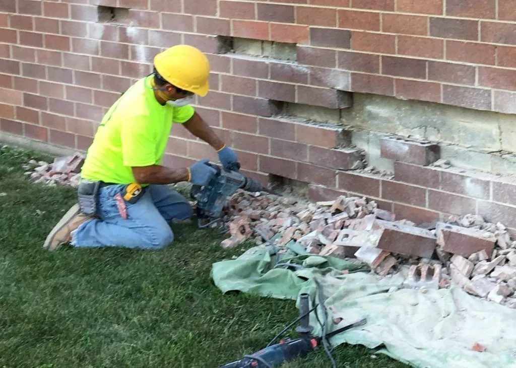 A worker in a neon yellow shirt and hard hat uses a power drill on a brick wall, creating a pile of rubble on the grass.