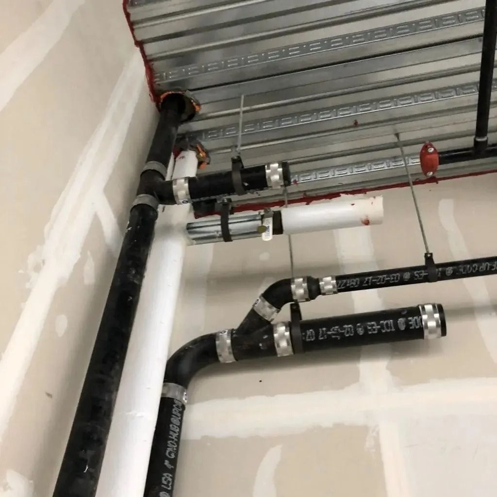 Black and white pipes are suspended from a corrugated metal ceiling and drywall in an unfinished room.