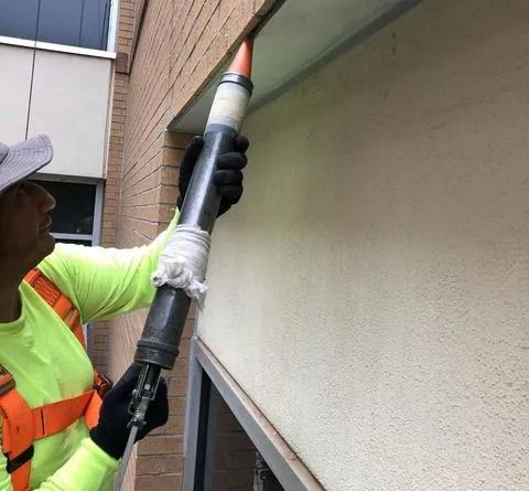 A person wearing high-visibility work gear applies sealant to a building gap with a caulking gun.