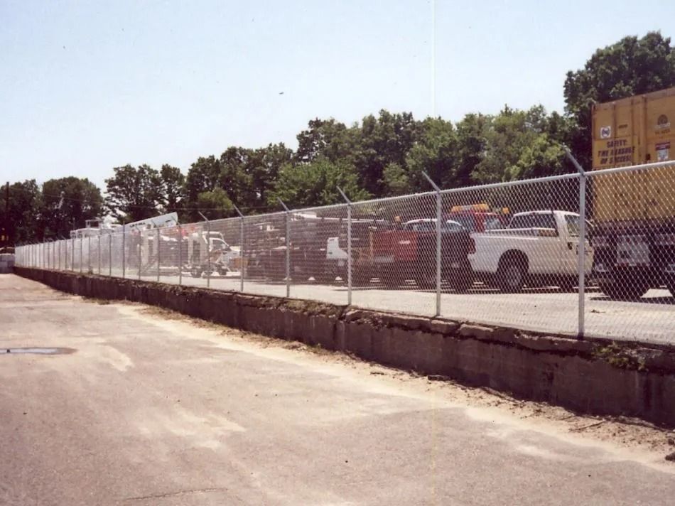 A chain-link fence topped with barbed wire runs along a concrete retaining wall bordering a fenced-in parking lot.