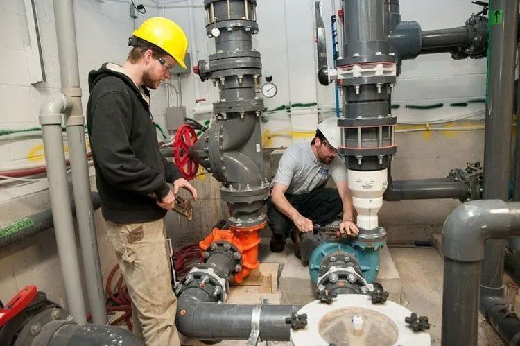 Two workers in hard hats and safety gear install or inspect large industrial pipes and pumps in a mechanical room.