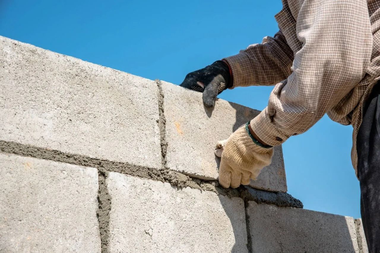 A construction worker in gloves places a concrete block onto a partially built wall against a bright blue sky.