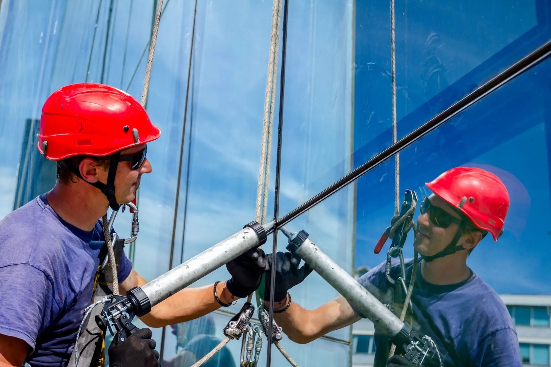 Two industrial workers in red helmets and blue shirts use sealant guns to repair the glass facade of a tall building.