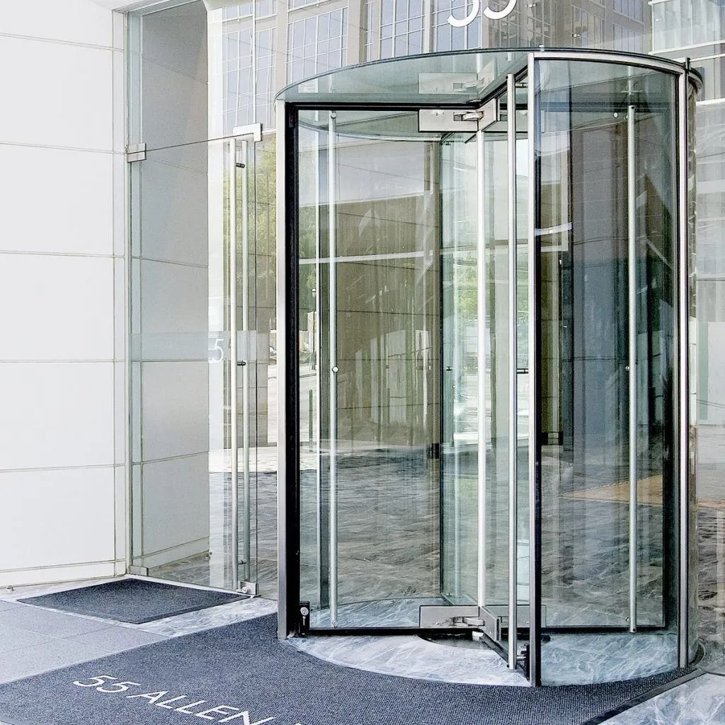 A modern glass revolving door at a building entrance featuring metal frames and grey floor mats.