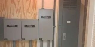 Three gray electrical disconnect boxes mounted on a plywood wall next to a tall, gray electrical service cabinet.