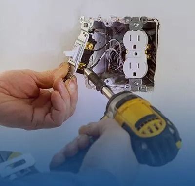 Hands use a power drill to secure a wall light switch next to an electrical outlet in a junction box.