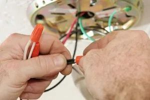 Hands connecting electrical wires with an orange wire nut for a ceiling fixture installation.