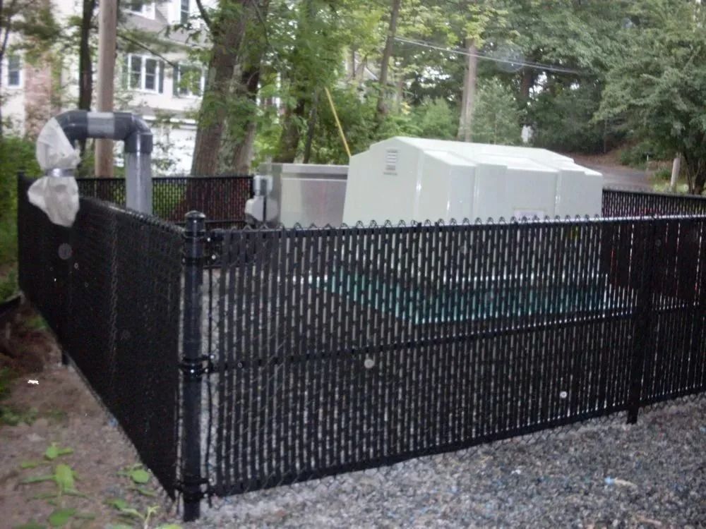 A black chain-link privacy fence surrounds a white utility enclosure and a grey pipe on a gravel surface outdoors.