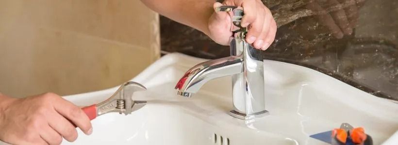 A person uses an adjustable wrench to repair a chrome bathroom faucet mounted on a white sink.