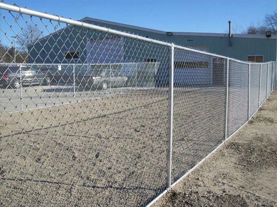 A chain-link fence encloses a gravel lot in front of a blue industrial building with garage doors.