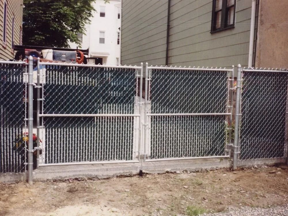 A double gate made of chain-link fencing with green privacy slats, installed in front of a house on a dirt lot.