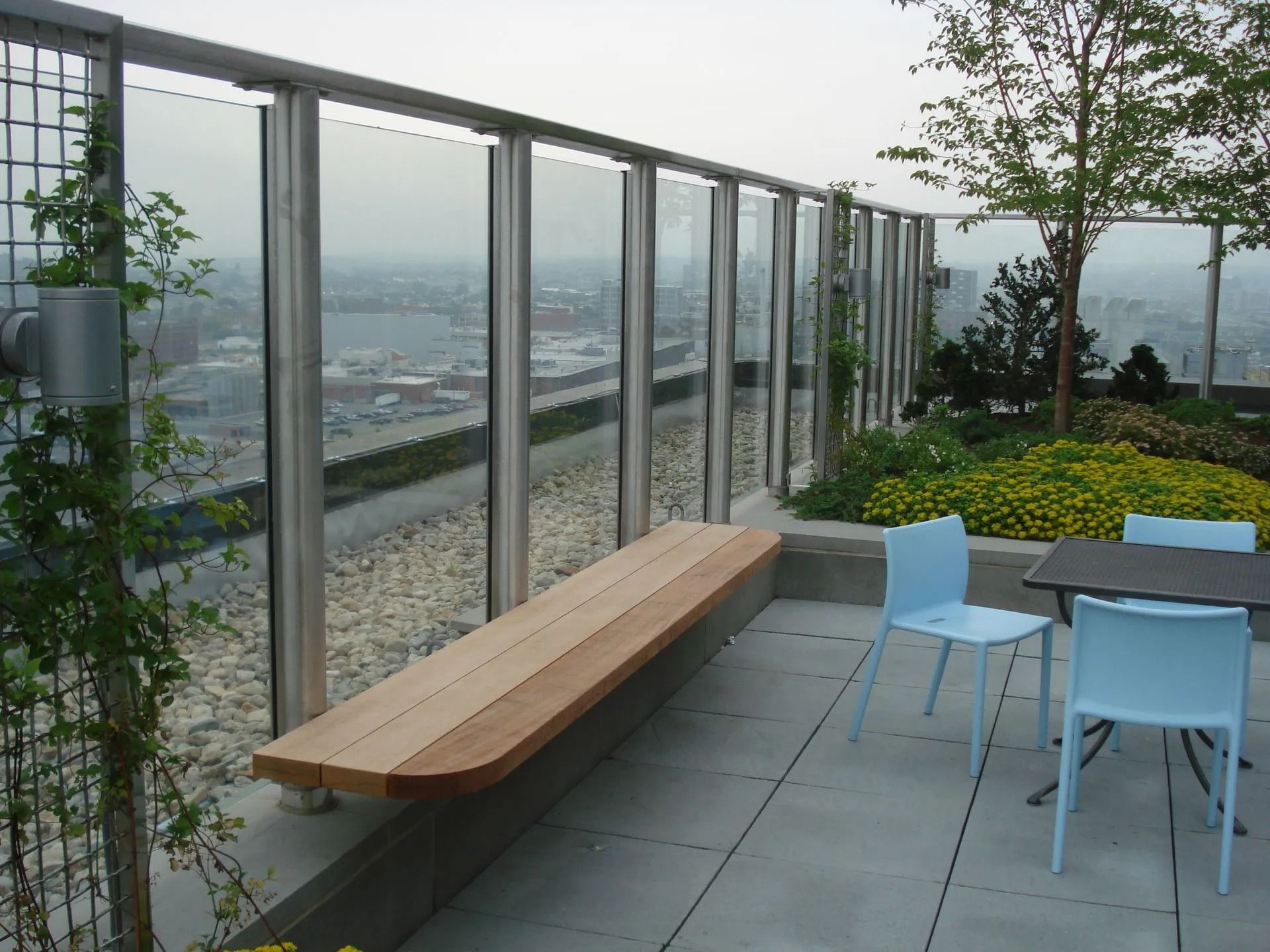 A rooftop terrace featuring a wooden bench against a glass railing overlooking a city, with chairs and potted plants.