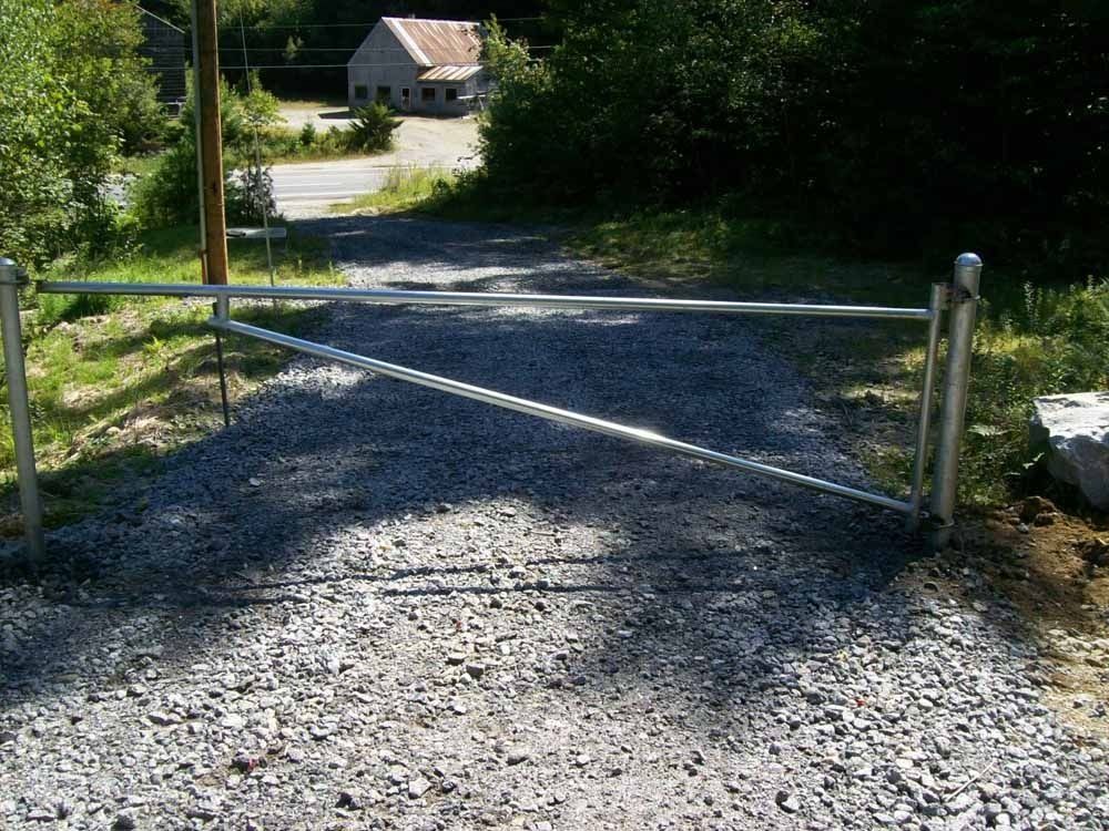 A gravel driveway blocked by a metal gate, with a small building visible in the background among trees.