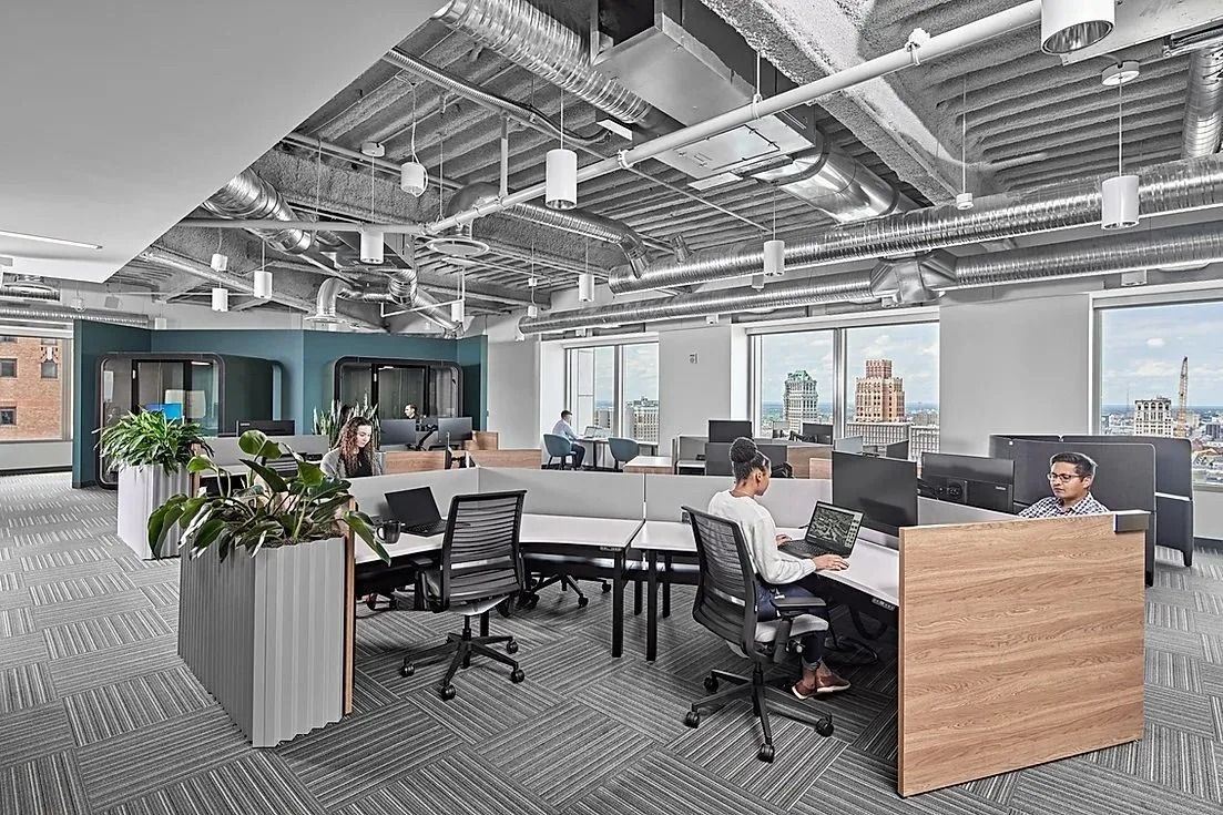 A modern, open-plan office with cubicle desks, industrial ceilings, and large windows overlooking a city skyline.
