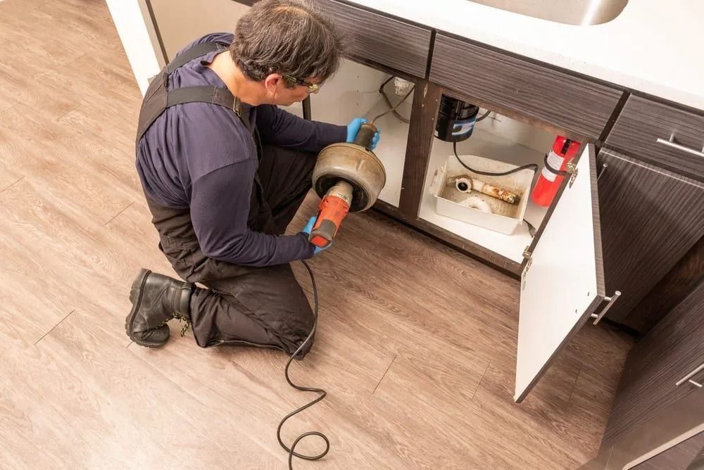 A plumber wearing dark overalls kneels on a wooden floor, using a drain snake to clear a pipe under a kitchen sink.