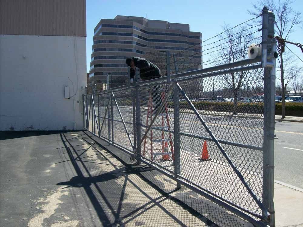 A person stands on a ladder, working on a chain-link fence with barbed wire in front of a building.