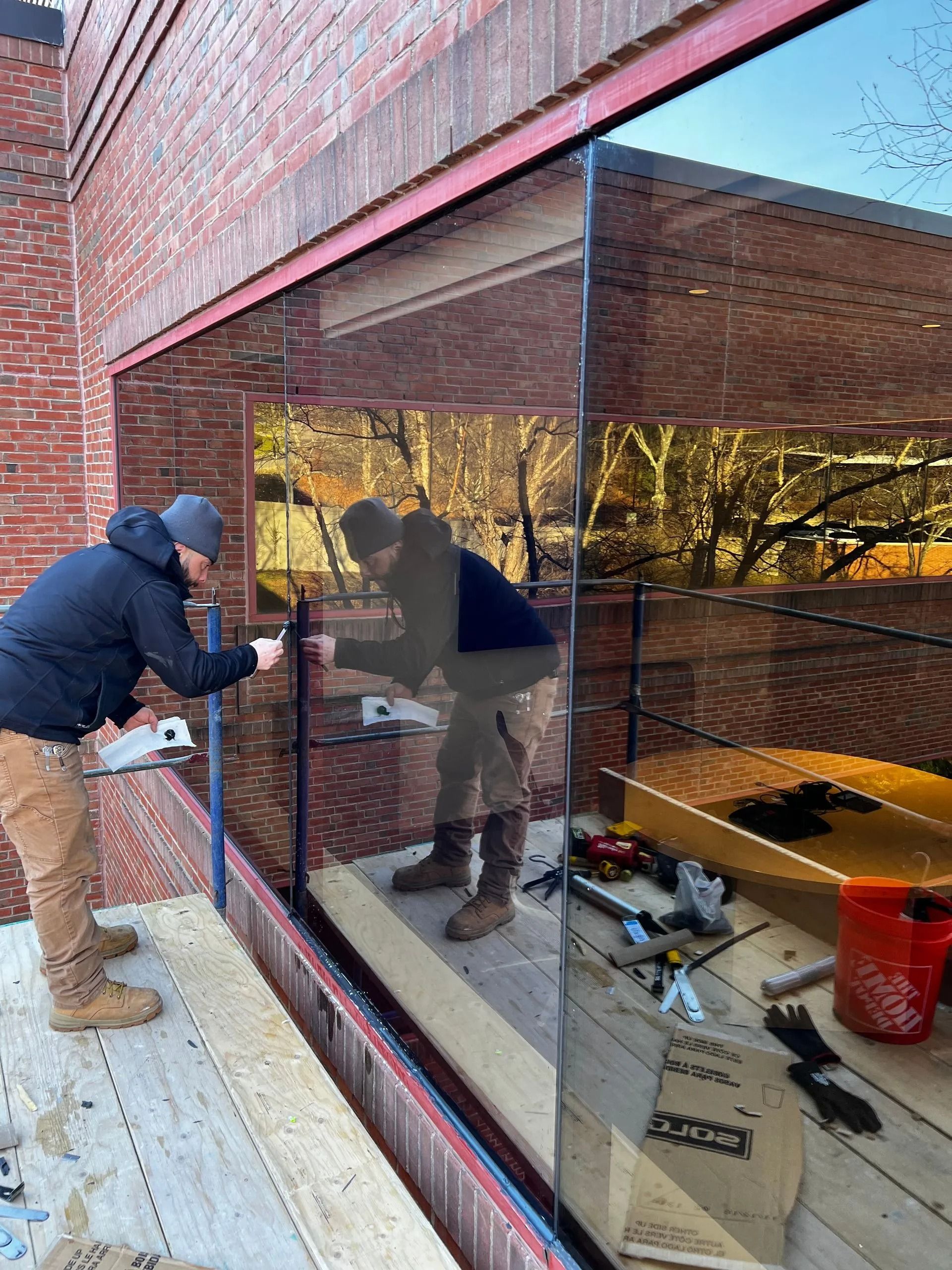 Two workers in hard hats and work gear install a large glass pane into a red brick building exterior on a scaffold.