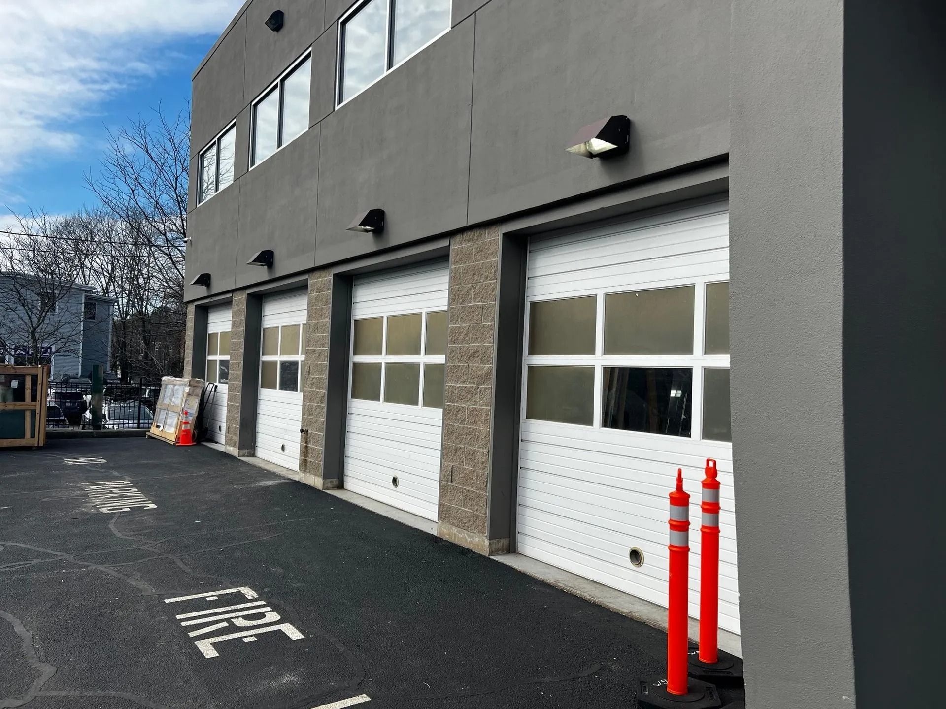 A grey, multi-story commercial building featuring three white garage doors with glass panels and red parking bollards.
