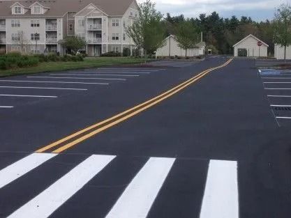 A parking lot with a freshly painted white crosswalk and a yellow double center line leading toward buildings.
